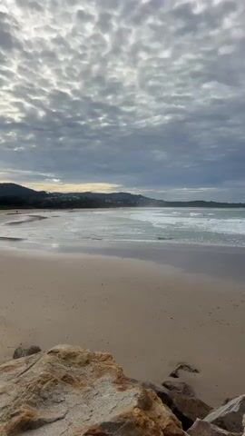 The video shows a vertical, wide-angle view of a beach on an overcast day. The foreground is dominated by large, tan-colored rocks. The middle ground features a sandy beach with a gentle slope towards the ocean, where waves are breaking. The ocean is a greenish-blue color and reflects the muted light from the sky. In the background, a low, tree-covered mountain range stretches across the horizon. The sky is filled with heavy, textured clouds in shades of gray and white, with hints of brighter light peeking through. There are no people or animals visible in the frame. The overall impression is one of a calm, natural coastal landscape under a cloudy sky.