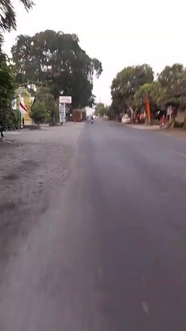 The video is a first-person perspective shot from a moving vehicle, likely a motorcycle, traveling down a paved road. The time of day appears to be late afternoon or early evening, with a bright sky and long shadows. The road is bordered by trees on both sides, and there are buildings and signs visible on the left side of the road. In the distance, several vehicles, including trucks and motorcycles, are also on the road. A motorcycle with a rider is visible in the foreground on the right side of the frame, also traveling in the same direction. The overall scene depicts a rural or suburban roadside environment.