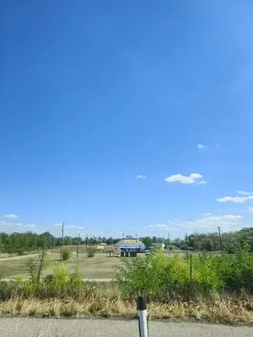 The video shows a bright, clear blue sky with a few wispy clouds. In the distance, there is a flat, grassy field with some trees and scattered structures. One prominent structure appears to be a building with a yellow roof. There are power lines visible in the background. The foreground shows dry grass and brush, and what looks like a road or path. The scene appears to be rural or suburban.