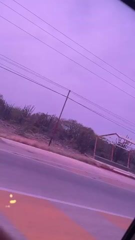 The video shows a street view from a moving vehicle, likely a car. The sky is a dull purple color.  A few sparse trees, cacti, and utility poles are visible along the side of the road, as well as structures that appear to be dilapidated or abandoned. The road itself has striped lane markings. Loud music plays in the background.