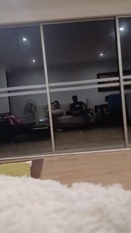 A person is sitting on a couch, holding a pillow and making a peace sign with their fingers. They are wearing a light-colored hooded sweatshirt and shorts, and white sneakers. Next to them, another person is sitting on the couch, looking down at their phone. They are wearing a dark t-shirt and jeans. They are both sitting on a striped couch with pillows. The background appears to be a living room with a large glass sliding door. Outside the door, there is some greenery and lights. The floor in front of the glass door is wooden.