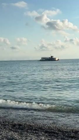 A medium shot captures a serene ocean scene under a partly cloudy sky. The foreground features a pebbled beach with small waves gently lapping at the shore, creating a foamy white crest. In the mid-ground, the calm blue water stretches out towards the horizon. A white passenger boat with multiple decks is visible sailing on the water, positioned slightly to the left of the frame's center. The boat appears to be in motion, its silhouette clear against the sea. The sky above is a light blue, dotted with fluffy white clouds that are illuminated by sunlight, suggesting it is daytime, likely late afternoon or early morning due to the soft light. The overall mood is peaceful and tranquil. No text is visible in the frame.
