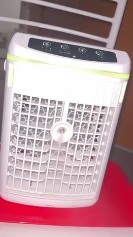 This video shows a close-up of a white portable air cooler with a honeycomb pattern grill. The air cooler has a control panel on top with several buttons and indicator lights, and it appears to be cycling through different colored LED lights. The air cooler is resting on a red surface, possibly a chair or table, and a white object, likely part of a chair or stand, is visible in the background. The video is very short, with the lighting changing subtly throughout.