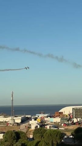 The video shows an aerial view of an outdoor event with two small planes performing aerobatics in the sky, leaving smoke trails. Below the planes, there is a large crowd of people gathered, with several temporary structures and stages set up, suggesting a festival or airshow. In the background, a body of water can be seen, possibly a bay or ocean, with a distant shoreline and buildings. The sky is clear and blue. The overall scene depicts an active outdoor event with an air show component.