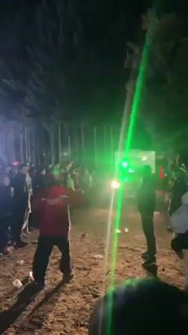 The video shows a nighttime outdoor gathering of people in a wooded area. A white vehicle with headlights on is slowly moving through the crowd. A stage with bright lights is visible in the background. The crowd is a mix of people wearing various clothing, including a person in a red jacket and another in a white jacket. The ground is uneven, appearing to be dirt or mud. There are trees lining the sides of the scene. Music is playing loudly.