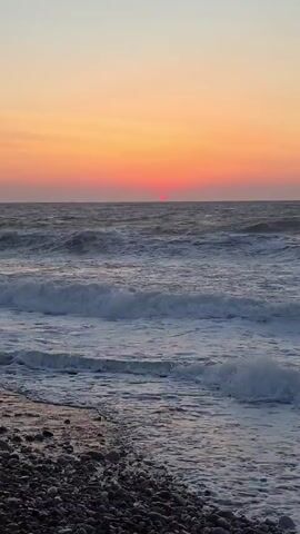 The video shows a sunset over a choppy ocean. The sun is a small, bright red orb just above the horizon. The sky is a gradient of orange and pink. Waves are crashing on a pebble beach. The water is a dark blue-gray and the waves are white-capped. There are no people or objects visible in the video.