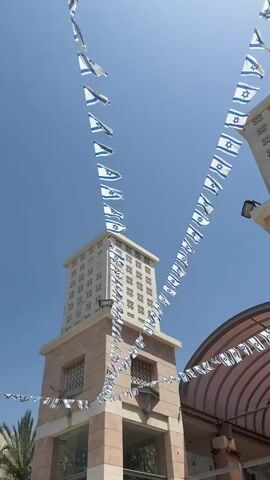 The video shows a clear blue sky, with several buildings adorned with strings of Israeli flags. The flags are small and rectangular, displaying the Star of David in blue on a white background. The buildings appear to be constructed from light-colored stone or stucco. The central building features a tower with a repeating pattern of square openings. A curved awning is visible in the foreground, also decorated with the Israeli flags. There are no people visible in the frame. The flags are gently moving in the breeze.