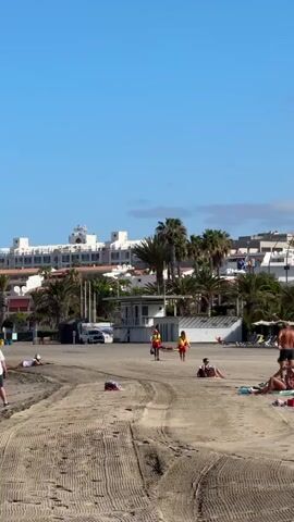 This video captures a sunny day at a beach in Los Cristianos, Spain. The scene is set on a sandy shore with the ocean just out of frame. In the background, a large, multi-story white building with many windows lines the coast, suggesting a resort or hotel complex. Palm trees are scattered throughout the area, adding to the tropical ambiance.

Several people are visible on the beach. Two individuals, dressed in red and yellow, appear to be lifeguards, walking across the sand. Other beachgoers are scattered, with some lying down, likely sunbathing, and others sitting. A white vehicle, possibly a van or small bus, is parked near a small building with a white roof, which could be a beach facility or lifeguard station. The sand itself shows tire tracks, indicating recent activity, perhaps from maintenance vehicles.

The sky is a clear, bright blue, with no visible clouds, suggesting fair weather. The bright sunlight and shadows cast by the people and objects indicate it is midday or early afternoon. The overall mood appears relaxed and leisurely, typical of a vacation destination. No visible text is present in the video.