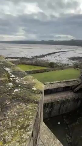 The camera pans right from atop a stone castle wall, which is covered in green moss. Below the wall is a square pit with irregular stone walls and visible shadows. The castle itself is made of gray stone and includes chimneys and roofs. Beyond the castle wall is a long, green field that appears to have a few sheep in it. Beyond the field is a view of a mud flat which extends to the edge of the frame. A green treeline is visible on the opposite edge of the flat. The sky is overcast and gray.