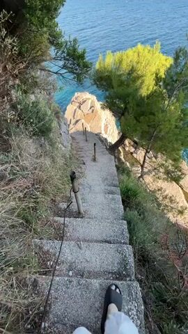 The video shows a set of concrete stairs descending towards a rocky cliff overlooking a blue sea. The stairs have metal poles along the sides. The surrounding area is covered with vegetation, including trees and dry grass. In the distance, islands can be seen on the horizon. The sky is clear and blue with a few scattered clouds. The camera moves, initially showing a first-person view of someone walking down the stairs, then pans to the left, revealing the expansive ocean view.