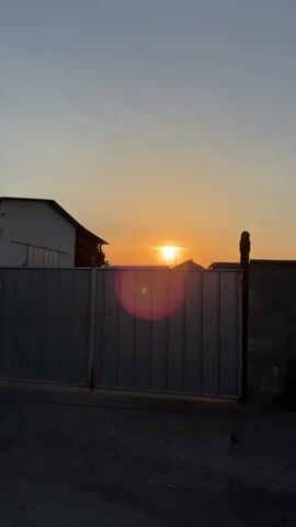 The video shows a sunset behind a large, grey metal gate. The sun is a bright orange orb, partially obscured by a building or structure. Lens flare appears on the left side of the frame. The sky transitions from a pale yellow near the horizon to a light blue higher up. To the left of the gate, a white building with a dark roof is visible. To the right of the gate, a brick wall and a dark vertical post are present. The ground in the foreground appears to be asphalt. The overall scene depicts an outdoor environment during sunset.