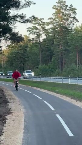 A woman in a red hoodie and denim shorts rides a bicycle on a paved path next to a road. She is smiling and waves at the camera as cars drive by on the road. The path is lined with trees and the road is separated from the path by a guardrail.
