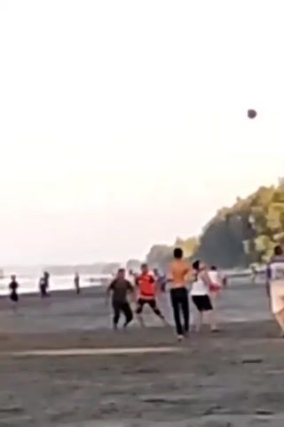 A group of people are playing soccer on a dark sand beach. The water is visible in the background, with waves breaking on the shore. The sky is overcast and the overall lighting suggests it is late afternoon or early evening. The people are wearing casual clothing, with some of them shirtless. There are other people in the background, some of whom appear to be watching the game. The scene is blurry due to the low resolution of the video.