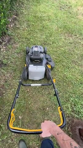 The camera films over the shoulder of a person operating a black and grey lawnmower with a yellow handle. The person is wearing blue pants and green boots. The lawnmower is on a lawn with a small bush and a patio in the background. The person is holding the lawnmower handle and the machine is moving forward.