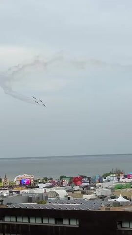 An aerial view shows four planes flying in formation, leaving smoke trails in the sky. Below, a city or town is visible with buildings, tents, and what appears to be a festival or event setup with stages and colorful banners. The ocean is visible in the background under a cloudy sky.