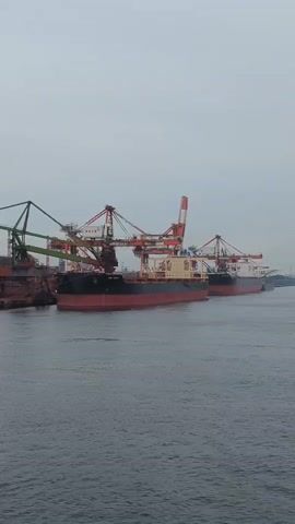 The video shows a large industrial port with several cargo ships docked. Cranes are positioned over the ships, likely for loading or unloading cargo. The port is surrounded by industrial facilities, including smokestacks emitting plumes of smoke. The water appears calm, and the sky is overcast. The scene suggests ongoing commercial activity at a busy shipping hub.