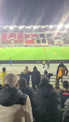 The video shows a soccer game at a stadium. The camera is behind a crowd of spectators. A man in a checkered hat is in the foreground. Many spectators are wearing jackets. In the background, players in red and yellow uniforms are on the green soccer field. The stadium is well-lit and there are fans in the stands. The field is encircled by a running track. Some spectators are standing on the track behind a railing.