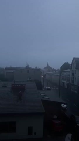 A view from above a town at dusk shows a street with a few cars. Buildings line the street, some of which are residential and others appear to be commercial. The sky is overcast and hazy, suggesting poor weather conditions. In the distance, a church spire rises above the rooftops. The scene is dark and the lighting is dim, indicating it is either early morning or late evening. The video captures the tranquil atmosphere of the town under a cloudy sky, with a hint of the impending storm.