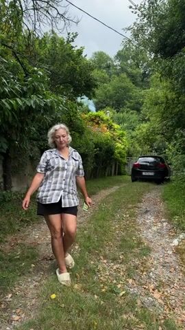 A woman with blonde hair, wearing a plaid shirt and black shorts, stands on a dirt path surrounded by greenery. She walks towards the camera, stops, and smiles. She then looks up and away before returning her gaze to the camera with a slight smile. A black car is parked on the path behind her.