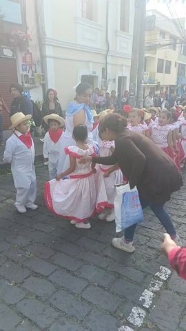 A group of children, dressed in traditional clothing, dances in a parade. The girls wear white blouses with red polka dots and red skirts with white polka dots, straw hats, and red flowers in their hair. The boys wear white pants and shirts with red scarves and straw hats. An adult woman in a light blue dress leads the group. Spectators watch from the sidewalks lined with buildings. Colorful banners hang overhead. The street is paved with cobblestones. One woman in jeans and a brown coat directs the children. A paper bag with the words “Juanito Popote” is visible in her hands. Music plays in the background.