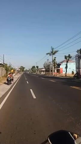 The video shows a highway view with a clear blue sky. The road is paved with white lines indicating lanes. On the left side of the road, there are sidewalks, trees, and buildings. On the right side, there are more trees, buildings, and a few cars. The video is filmed from a moving vehicle, likely a motorcycle, given the visible handlebars in the foreground. The scene appears to be a rural or suburban area during the daytime.