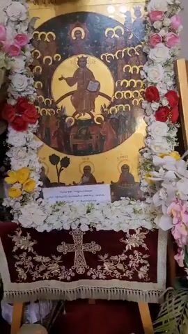 The video pans across the interior of an Orthodox church in Monagrouli, Cyprus. The scene is richly decorated with religious iconography and floral arrangements. In the foreground, a large, ornate icon of Christ Pantocrator is visible, surrounded by a golden halo and a multitude of smaller figures. The icon is adorned with white and pink artificial flowers, and a velvet cloth with a cross design is draped at its base. A piece of paper with Greek text is attached to the icon.

The camera then moves to reveal the church's interior, showcasing intricately carved wooden furniture, including large chairs and a pulpit. The walls are adorned with numerous icons depicting religious figures and scenes. A brass candelabra stands prominently on the red carpeted floor. A vase of white lilies and other flowers adds to the decorative elements. The lighting suggests it is daytime, and the overall atmosphere is one of solemnity and reverence. The absence of people suggests the video was recorded during a quiet time in the church, possibly before or after a service.