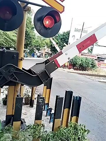The video captures a railway crossing in Sidoharjo, Indonesia, during what appears to be daytime under a cloudy sky. The red and white striped crossing gates are in the process of lowering, and the red flashing lights of the signal are actively blinking. A distinct warning bell sound is audible, signaling an approaching train. Several vehicles, including cars and motorcycles, are visible stopped on the road, waiting at the closed crossing. In the background, train tracks run parallel to the road, and various buildings are present, including a large billboard displaying the text "Idul Fitri." Trees and other foliage are seen lining parts of the road and tracks.