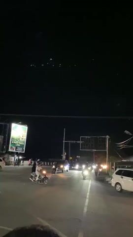 The video captures a nighttime scene on a street in Lambheu, Indonesia. The time of day is clearly indicated by the dark sky, illuminated only by streetlights, vehicle headlights, and a large, brightly lit billboard.

The subjects are a mix of people and vehicles. Several motorcycles are visible, some carrying passengers. A few cars are also present, their headlights cutting through the darkness. On the left side of the street, a prominent billboard displays an advertisement with text that includes "A MAN TASTE" and an image of what appears to be food or a beverage. The setting is urban, with buildings and traffic lights lining the street, suggesting a relatively busy road, possibly at an intersection.

The activity taking place is the movement of traffic. Motorcycles and cars are navigating the road, their lights creating streaks and reflections on the wet-looking asphalt, hinting at recent rain or a humid atmosphere. The overall mood is that of a typical evening commute or late-night activity in a tropical urban environment. The lights create a vibrant, somewhat hazy, atmosphere against the deep black of the night sky. The scene feels dynamic due to the constant motion of the vehicles.