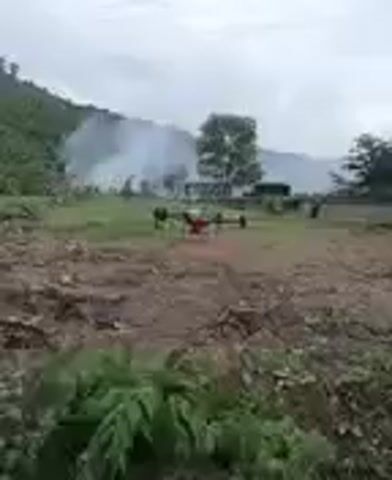 A quadcopter drone hovers in the foreground of a rural, outdoor scene. The drone is orange and black and has four propellers. In the mid-ground, there is a patch of green grass and several trees. Smoke billows from the left side of the mid-ground, obscuring some of the scenery. In the background, a forested hillside rises under an overcast sky. To the left of the frame, in the foreground, there are some green plants and brown dirt with scattered rocks. To the right, there is more brown dirt and sparse vegetation. There are some structures and what appear to be people in the mid-ground area.