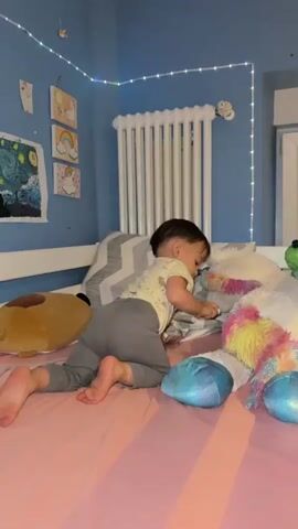 A young child with dark hair is crawling on a pink bed in a room with blue walls. The child is wearing a yellow shirt with a white pattern and gray pants. The child is holding a small object, possibly a toy or a marker, and appears to be examining it. The bed is covered with a pink sheet and several stuffed animals, including a large white unicorn with a colorful mane and a brown bear. There are also pillows on the bed, one with a gray and white chevron pattern. In the background, a white radiator is mounted on the wall, and a string of white fairy lights is draped along the top of the wall. Several framed pictures are hanging on the wall, including one that resembles Van Gogh's "Starry Night" and others with images of rainbows and unicorns.