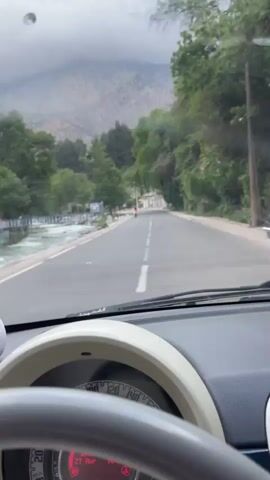 The video captures a drive through a mountainous landscape, initially from the driver's perspective and then from the passenger side window.

**Subjects:** The scene features a paved road running parallel to a fast-flowing river. Lush green trees line the riverbank, and rugged mountains, partially shrouded in clouds, rise in the background. A building with a colorful facade and a small bridge spans the river at one point. Inside the car, a steering wheel, dashboard, and a speedometer showing red illuminated numbers are visible. A white Pudu deer plush toy rests on the dashboard. The side rear-view mirror reflects the road behind, showing another car and the arm and shoulder of a person, likely a passenger.

**Activity or event taking place:** The video shows a car driving along a scenic road. The initial shot is from inside the vehicle, looking out the front windshield, and then transitions to a view from the passenger's side window, capturing the passing scenery.

**Notable details:**
- **Time of day:** Daytime, indicated by ambient light, but the sky is heavily overcast.
- **Weather:** Cloudy and grey, with mountain peaks disappearing into the low-hanging clouds.
- **Location cues:** The combination of mountains, a river, and the specific architecture of the building and bridge are consistent with the provided location context of Asgaour, Morocco.
- **Emotions:** The scene evokes a sense of travel and appreciation for natural scenery.
- **Visible text:** No legible text is visible in the video.