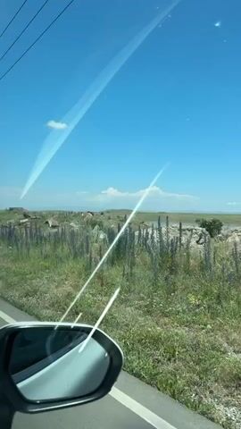 This video, filmed from inside a car, captures a scenic drive through Nor Kyanq, Armenia. The view outside the car window reveals a vast, open landscape under a bright blue sky with scattered white clouds. The sun is shining, suggesting a clear, warm day, likely daytime given the brightness.

The foreground of the video is dominated by the car's side mirror and the side of the windshield, which shows streaks of light or smudges. Beyond the car, the landscape is characterized by rolling hills covered in green grass and dotted with wildflowers, predominantly purple lupines and yellow blossoms. In the mid-ground, a body of water, likely a lake or reservoir, stretches across the horizon. Several vehicles are visible near the water's edge, suggesting a popular spot for recreation or rest. Some structures, possibly ruins or small buildings, are visible on a distant hill.

The overall impression is one of a peaceful, natural setting being enjoyed during a road trip. The activity is simply driving and observing the scenery. There is no discernible text in the video, and no people are clearly identifiable. The atmosphere is serene and expansive.