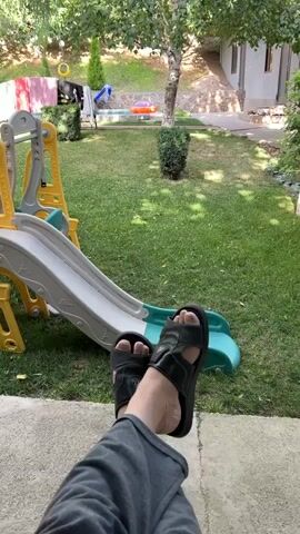 A person's feet wearing black sandals are visible in the foreground, with the camera angled upwards to capture a backyard scene. The backyard features a green lawn, a children's slide set, trees, and a building in the background. Clothes are hanging on a line in the distance.