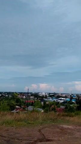 The video captures a panoramic view of a small town under a cloudy sky. The camera is positioned slightly elevated, looking down upon a cluster of houses and buildings with a mosque visible in the distance. The foreground is characterized by a grassy slope with patches of exposed soil. There are powerlines and utility poles scattered throughout the town, contributing to its visual landscape. The overall impression is of a serene, small town setting.