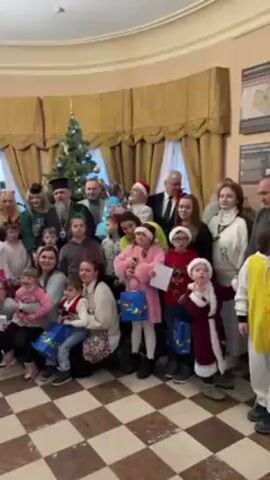 A group of adults and children are posing for a picture in front of a decorated Christmas tree. There are two priests in the group, and some of the children are wearing Santa hats or Christmas-themed clothing. Many of the children are holding gift bags. The room has a checkerboard floor and curtains in the background. A man is heard speaking in the background.