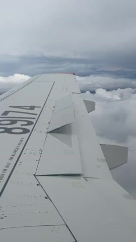 This is a first-person perspective video taken from an airplane window. The main subject is the wing of the aircraft, which is white with black lettering and various mechanical components. The wing is positioned to the left of the frame, extending diagonally across the image. There are visible rivets and panels on the wing's surface, as well as a section of the aircraft's fuselage on the far right.

The background is dominated by a cloudy sky. Thick, white clouds fill the lower half of the frame, with lighter, more dispersed clouds and a grey, overcast sky above. In the distance, faint outlines of mountains can be seen through the clouds, suggesting a mountainous or hilly landscape below.

The visible text on the wing reads "8974" in large, bold black numbers. Below this, there is smaller text that is difficult to read clearly due to its size and resolution, but it appears to be a set of instructions or warnings, possibly related to aircraft operation. One line clearly visible reads "DO NOT WALK OUTSIDE THE WING".

The time of day appears to be daytime, but the overcast weather conditions create a diffused, somewhat dim light. The overall mood is one of travel and observation, with the focus on the mechanics of flight and the vastness of the sky. There are no people or other objects visible in the frame besides the aircraft wing and the clouds. The location context provided is Murmashi, Russia, which hints at the geographical area from which this aerial view is captured.