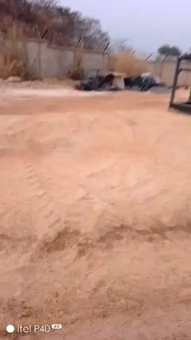 The video shows a wide, open dirt lot, possibly a construction site or industrial yard. The ground is reddish-brown and shows tire tracks. A large, dark-colored machine with a wheel is visible to the left. In the background, a row of stacked shipping containers in various colors are visible. A few palm trees and a pile of earth are visible further back. The sky is overcast and the lighting is dim. The video is shaky, indicating it was taken by hand. The lower-left corner of the video displays the text "itel P40 4G".