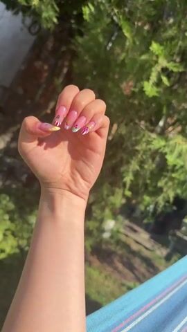 The media file is a close-up, vertical shot of a person's left hand and forearm, held up against a blurred background of greenery and a bright sky. The hand is clenched into a fist, with the palm facing the viewer. The subject's nails are long and almond-shaped, painted with a pink base and adorned with colorful, detailed designs of various fruits, including what appear to be strawberries, blueberries, and possibly grapes or cherries. Some nails also feature white tips or abstract colorful patterns. The skin tone is fair, and the lighting suggests it is daytime, likely sunny, given the bright highlights on the arm and the clear, well-lit appearance of the nails. The greenery in the background, which is out of focus, gives the impression of an outdoor setting, possibly a garden or park. The lower right corner of the frame shows a sliver of a blue and white striped fabric, hinting at a hammock or similar casual seating. The overall mood is vibrant and cheerful, likely conveying a sense of fun and personal style, perhaps showcasing newly done nail art. There is no visible text in the media file. The location context is Kameno, Bulgaria.