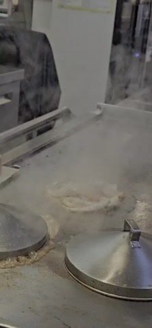 A person is using tongs to stir fry vegetables and seafood on a flat top grill. There is a lot of steam coming off the grill. There are two lids with handles on the edge of the grill. The person is wearing a bracelet on their left wrist. Another person can be seen in the background.