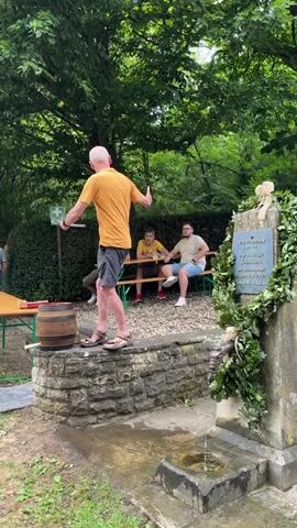 A man in an orange t-shirt and shorts stands on a stone structure, holding a paper and gesturing with his left hand. He appears to be addressing a small crowd gathered outdoors. To his right, a stone fountain with a wreath of greenery is visible, with water flowing from a spout into a basin. Behind the man, several people are seated on a bench, and others are standing, some clapping. Further back, a large banner with a coat of arms is displayed, attached to a vehicle.

The scene suggests an outdoor gathering or celebration, possibly an anniversary or festival, given the presence of a banner and a decorated fountain. The lush green trees and foliage indicate a daytime setting, likely during warmer weather. The expressions and actions of the people suggest a positive and engaged atmosphere.

Visible text on the stone monument reads: "Zur Erinnerung an/ 50-jährige Jubiläum/ des Brunnenfestes/ Meisenheim". This translates to "In remembrance of/ 50th anniversary/ of the Fountain Festival/ Meisenheim." This confirms the event is a 50th-anniversary celebration of a fountain festival in Meisenheim, Germany. The overall mood appears festive and community-oriented.