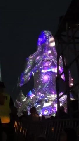 A night-time outdoor event features a massive projection of a female figure onto a dark surface. The projection depicts the figure covered in swirling cosmic patterns of purple, blue, and white, with glowing stars and nebulae. The figure has long, flowing hair and her hands are clasped over her chest. Below the projection, a crowd of silhouetted people are visible, gathered behind a metal barrier. The scene appears to be a concert or festival, with stage lighting and structures visible in the background. A security guard in a yellow vest is partially visible on the left side of the frame.