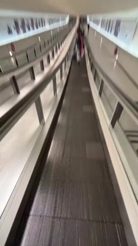 A camera travels along a moving walkway in an indoor, airport-like setting. Parallel walkways are to either side, with safety rails and advertisements visible. Several people with luggage are ahead on the same walkway, moving in the same direction. The walkway leads to an escalator. The lighting is bright and uniform.
