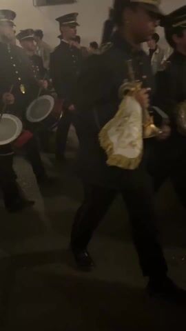 A group of men in black uniforms and peaked caps are walking and playing drums. The drums are white with the word "Cordoba" printed on them. The men are marching in a line, and there are other people in the background. The environment appears to be an outdoor street at night.