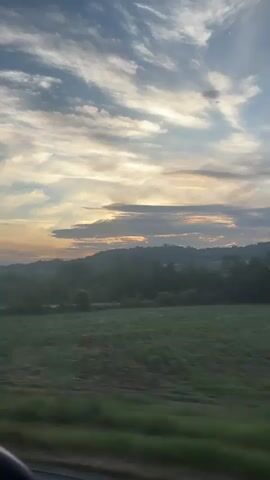 A person is traveling in a vehicle, looking out the window. The view outside the window shows a landscape with fields and trees, and a distant hill range. The sky is visible, with scattered clouds illuminated by the setting sun. The sun is partially obscured by a layer of clouds, creating a soft glow. The view is slightly blurred due to the motion of the vehicle.