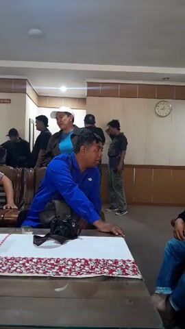 The video shows a group of people in what appears to be a room with wood-paneled walls and a clock on the wall. In the foreground, a man in a blue shirt and a white cap is seated at a table with a camera in front of him. He is looking to his right. Behind him, several other people are standing and talking. To the left, a person in a black shirt is partially visible. The overall lighting is dim.