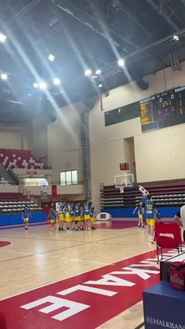 This video captures a basketball game taking place indoors in a gymnasium. The primary subjects are young male athletes, identifiable by their sports attire. They are engaged in a basketball game, dribbling, passing, and shooting the ball. The setting is a well-lit gymnasium with bleachers for spectators and a polished wooden court marked with standard basketball lines. The overall mood appears to be one of active competition and youthful energy.

The video's visual cues, such as the lighting and the presence of indoor facilities, suggest that the time of day is irrelevant for this indoor event. No external weather conditions are visible.

Several objects are prominent in the scene: basketballs, hoops with backboards, and scoreboards. The scoreboards display game-related information, including time and scores.

Visible text includes "BULGARISTAN" on a scoreboard, and banners or signs that appear to be related to sports development programs. Additionally, on the court's perimeter, the word "MARAKE" is prominently displayed in large letters. The presence of national flags, including one that appears to be the flag of North Macedonia, and portraits of important figures suggest this might be an international youth development event. The overall context points to a sporting event in Çanakkale Merkez, Turkey.