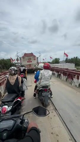 The video shows a convoy of motorcycles and a large truck on a bridge with red railings. Indonesian flags are flying on poles along the bridge. The sky is cloudy. Several people are riding on the motorcycles, some wearing helmets and headscarves. The truck appears to be a tanker truck. The motorcycles are moving in the same direction. The scene appears to be a busy crossing point.