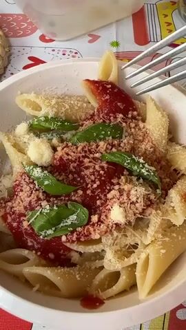 A close-up shot shows a white bowl filled with penne pasta topped with marinara sauce, fresh basil leaves, and grated Parmesan cheese. A fork is actively stirring the pasta, lifting a portion of it as if to take a bite. The scene is set against a colorful tablecloth adorned with heart patterns.