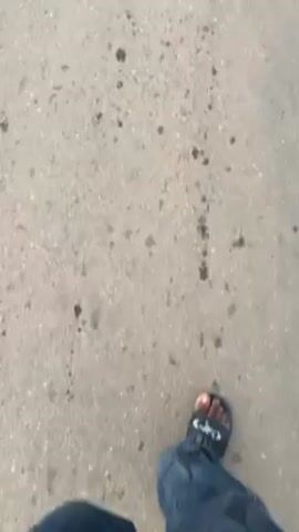 The video shows a first-person perspective of someone walking down a paved road. Initially, the camera focuses on the person's feet, shod in a graphic print shoe. As the video progresses, other people come into view, also walking along the road, wearing sandals or slide shoes. The camera then pans up to show the road ahead, revealing a long, straight stretch lined with green vegetation and palm trees. The sky is overcast and gray. The person in the video is wearing a plaid shirt and carrying what appears to be a bottle of water. Other people can be seen in the distance. The overall environment appears to be a rural or semi-rural area.