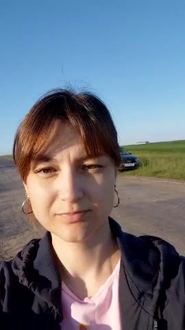 A woman with reddish-brown hair and bangs, wearing hoop earrings and a pink shirt under a dark jacket, smiles at the camera. She is standing outdoors on a dirt road next to a grassy field. The sky is blue and clear, and the sun is shining. The camera is tilted upwards, creating a slight fisheye effect. The woman appears to be moving, as the background is slightly blurred.